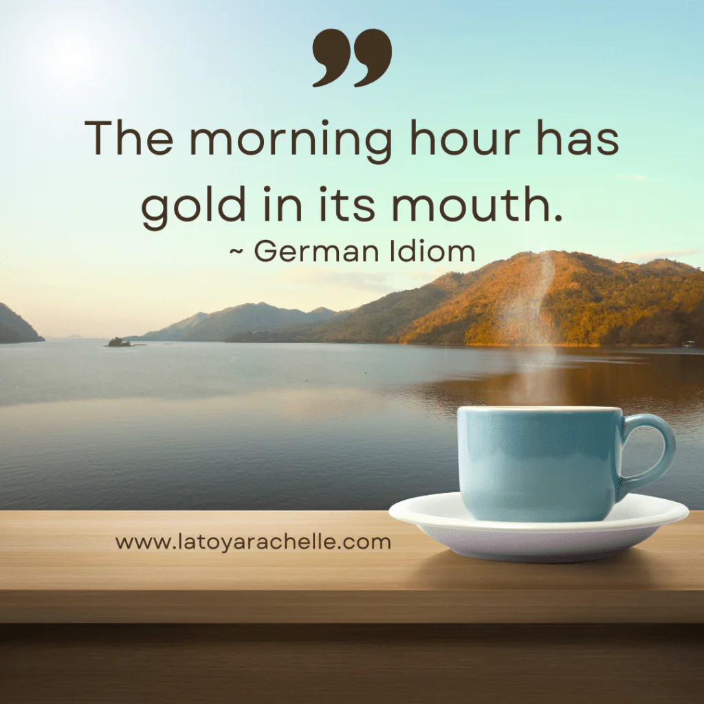 A blue coffee cup with steam sits on a wooden surface overlooking a serene lake surrounded by mountains under a clear sky. Text on the image reads, "The morning hour has gold in its mouth. ~ German Idiom." The website www.latoyarachelle.com is also displayed.
