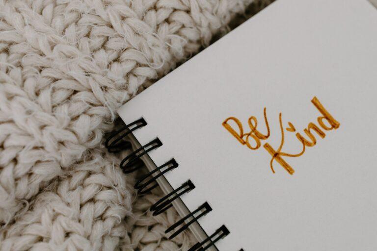 A close-up of a notepad with 'Be Kind' written in cursive gold ink, set against a cozy knitted background, capturing the essence of warmth and the message of the blog post 'Be Kind: 75 Kindness Affirmations to Help You Spread Kindness Daily'.