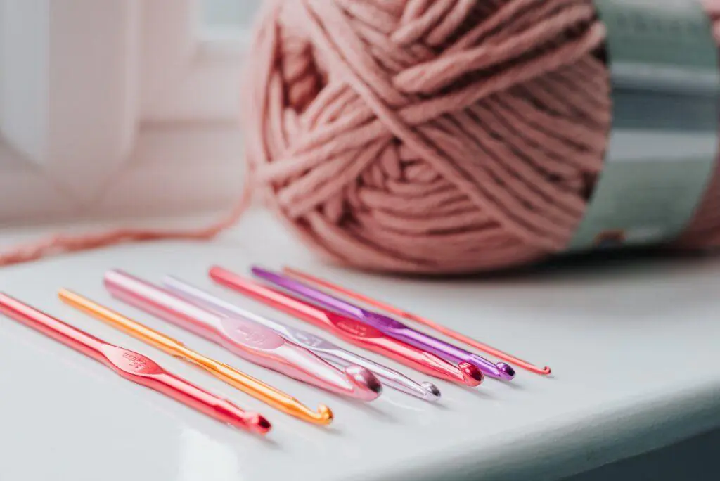 Colorful crochet hooks lined up in front of a ball of pink yarn on a white surface. This image highlights the tools and materials used in crocheting or knitting, relaxing and creative feminine hobbies.