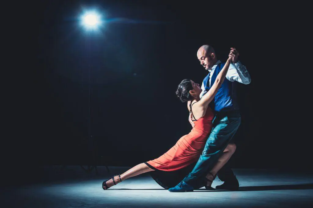 A couple performing an elegant ballroom dance, with the woman in a red dress and the man in a blue vest, under dramatic lighting. This image captures the passion and grace of ballroom dancing, a refined and expressive feminine hobby.