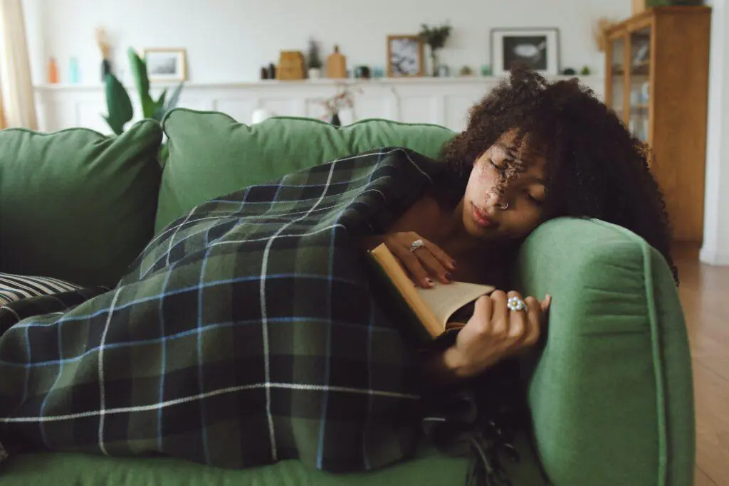 Woman relaxing on a green couch, wrapped in a plaid blanket, while reading a book. This image captures the serene and cozy atmosphere of reading, a beloved feminine hobby.