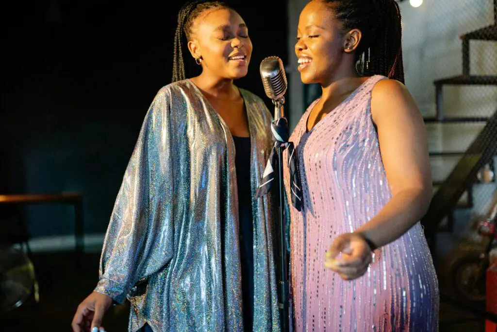 Two women joyfully singing into a vintage microphone, wearing sparkly outfits. This image captures the vibrant and expressive nature of singing, a feminine hobby.