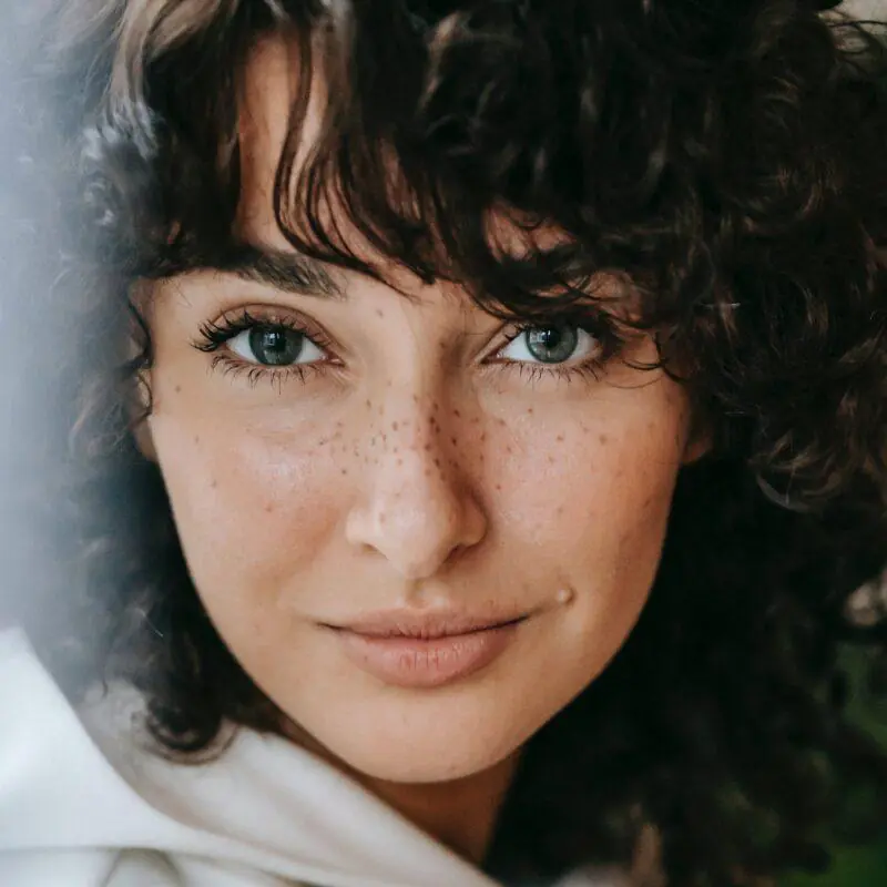 Close-up portrait of a woman with curly hair and freckles, gazing intently at the camera with green eyes. This image captures natural beauty and introspection, emphasizing the unique aspects of feminine energy.