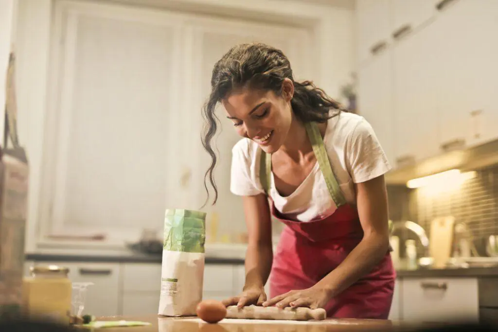 Woman smiling while rolling out dough on a kitchen counter, wearing a pink apron. This image illustrates the joy and creativity of cooking and baking, which is often considered a nurturing and feminine hobby.
