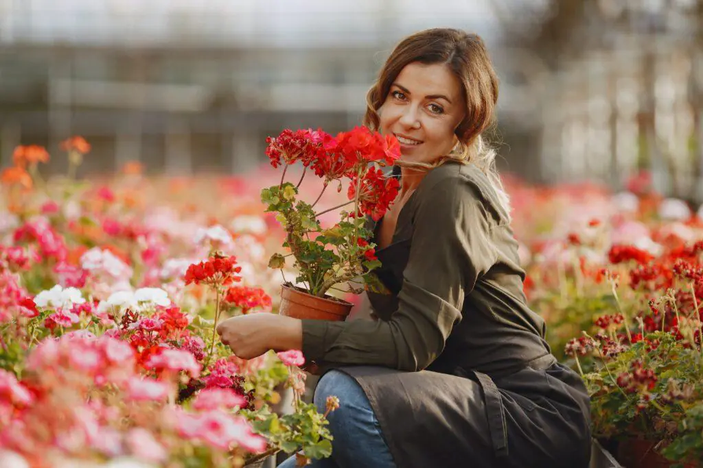 Woman smiling while holding a pot of vibrant red flowers in a lush garden, surrounded by an array of colorful blooms. This image highlights the joy and tranquility of gardening, a popular feminine hobby.