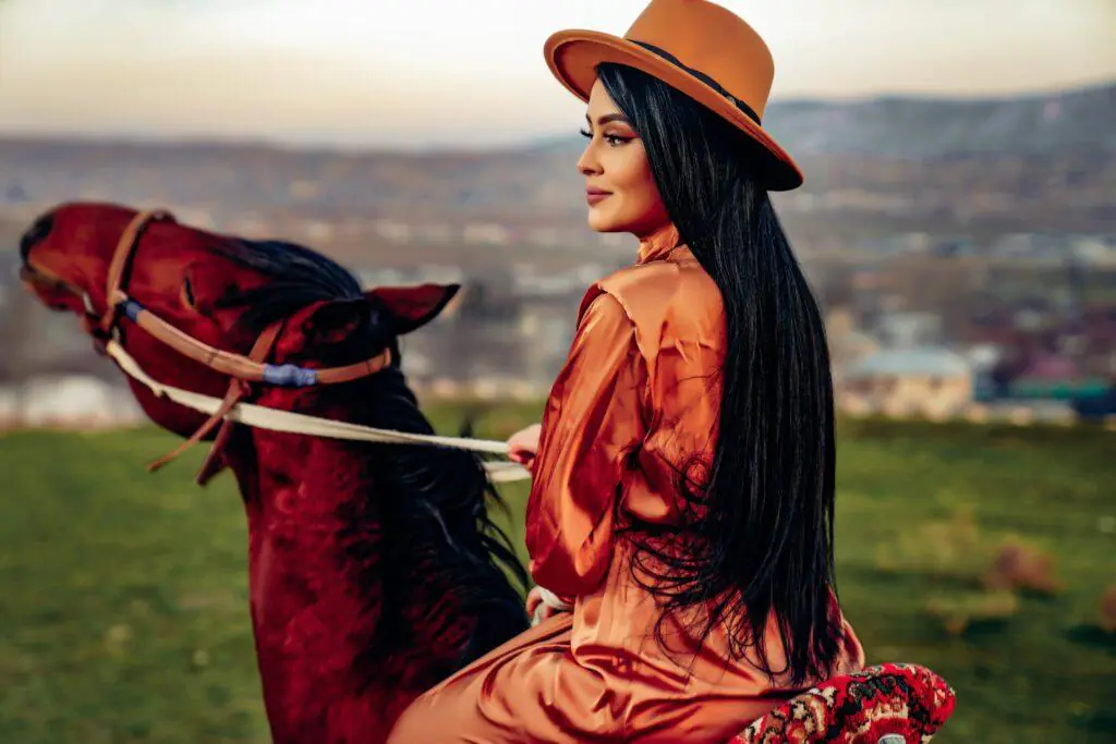 Feminine woman in an orange outfit and hat riding a horse, with a scenic landscape in the background. This image captures the elegance and adventurous spirit of horseback riding, a cherished feminine hobby.