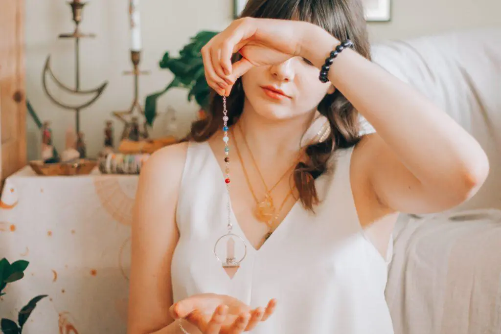 Woman holding a handmade pendulum with colorful beads, wearing a white top and multiple necklaces. This image depicts the intricate and artistic nature of jewelry making, a creative feminine hobby.