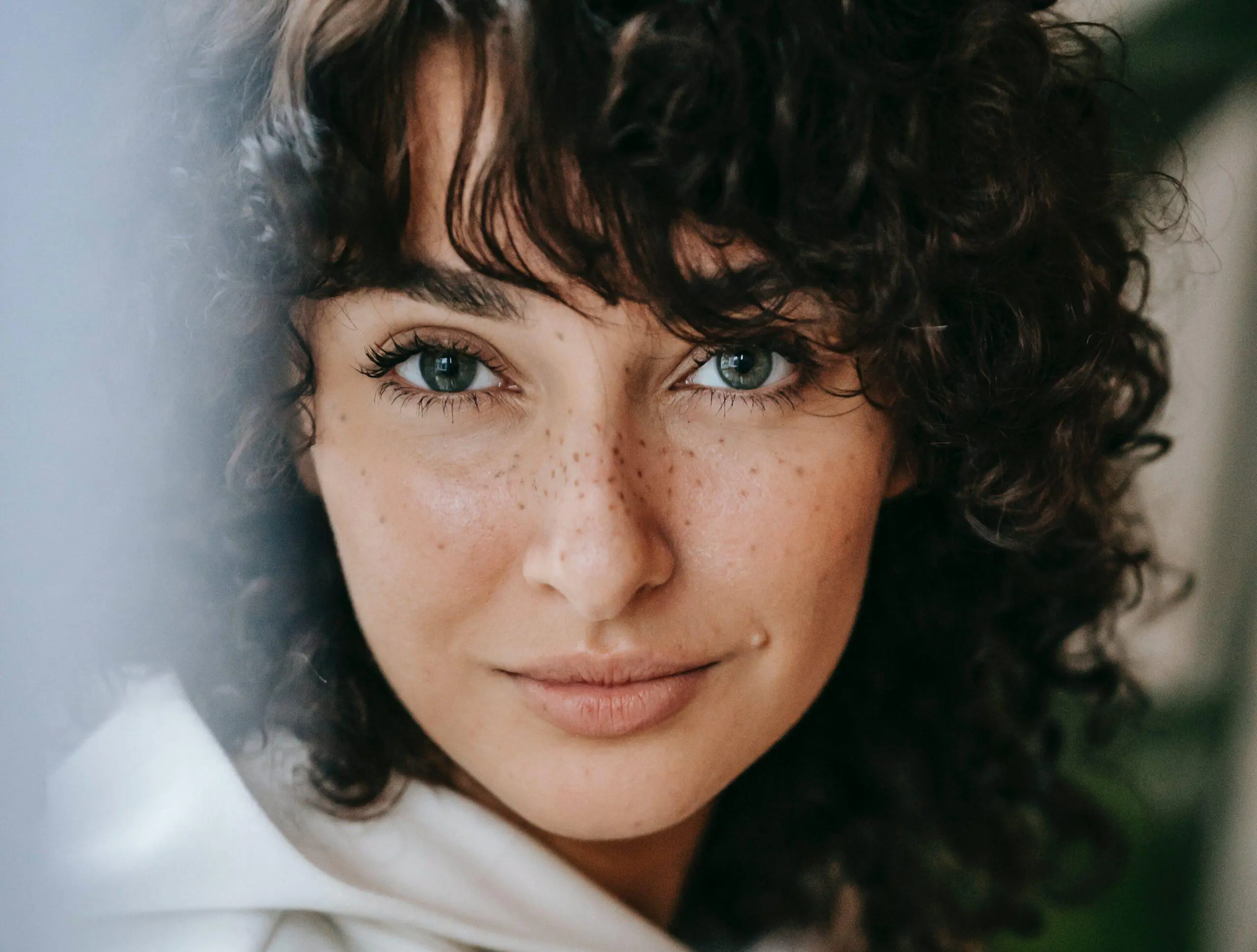 Close-up portrait of a woman with curly hair and freckles, gazing intently at the camera with green eyes. This image captures natural beauty and introspection, emphasizing the unique aspects of feminine energy.
