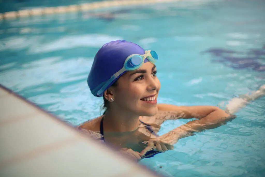 Woman smiling while leaning on the edge of a swimming pool, wearing a blue swim cap and goggles. This image represents the refreshing and invigorating hobby of swimming, which is often enjoyed as a feminine pastime.
