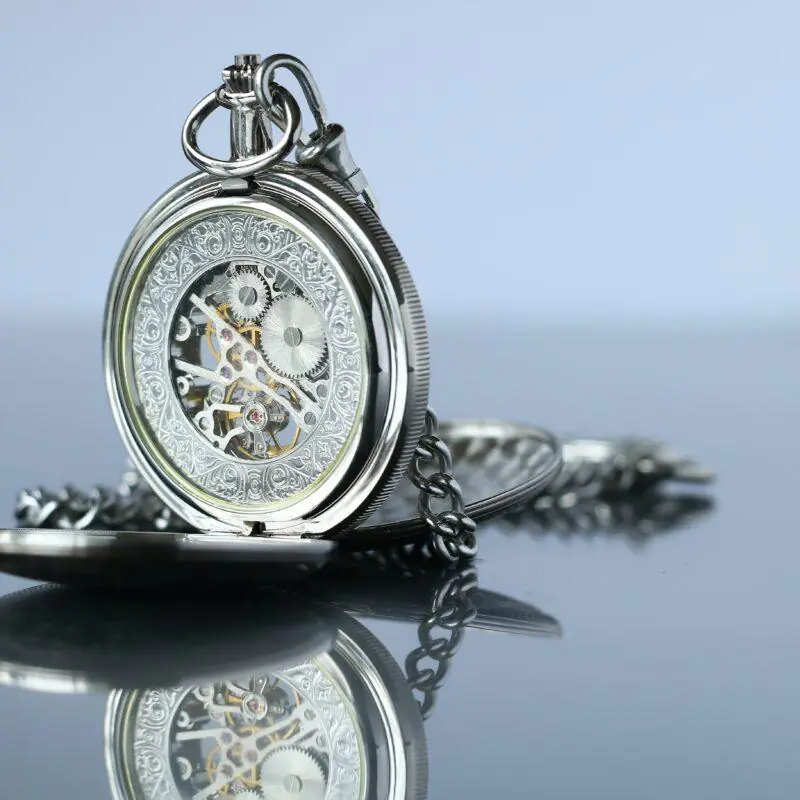 An ornate silver pocket watch with an intricately designed cover and visible inner gears lies open on a reflective surface. The image highlights the detailed craftsmanship, suitable for engraving short inspirational quotes.