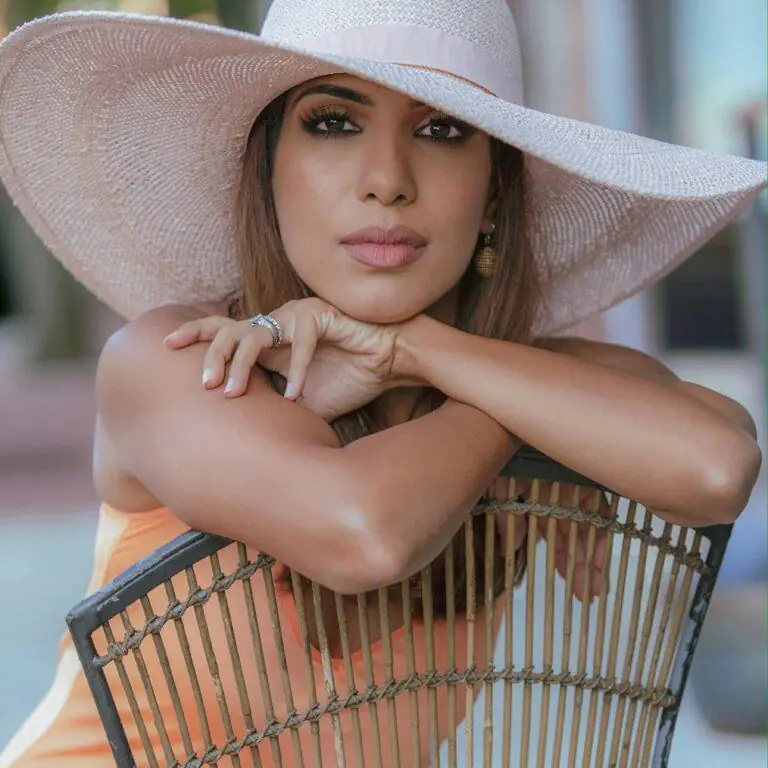 A woman wearing a wide-brimmed pink hat and subtle makeup leans on a wicker chair, looking directly at the camera. Her elegant pose and accessories highlight a refined and feminine style, making it ideal for a guide on how to be more feminine and ladylike.