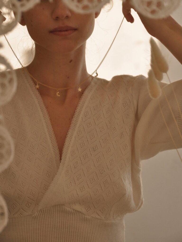 Close-up of a young woman partially concealed behind hanging lace decorations, holding a delicate gold necklace adorned with tiny star and moon charms. She wears a soft, textured cream sweater with a lace trim, capturing a serene and dreamy atmosphere.