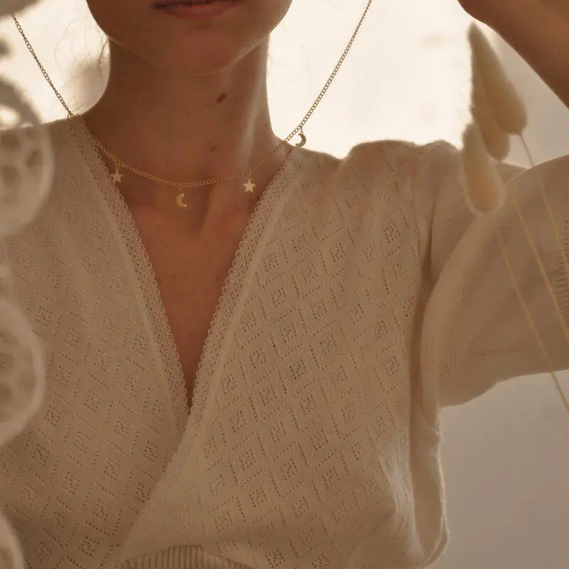 Close-up of a young woman partially concealed behind hanging lace decorations, holding a delicate gold necklace adorned with tiny star and moon charms. She wears a soft, textured cream sweater with a lace trim, capturing a serene and dreamy atmosphere.