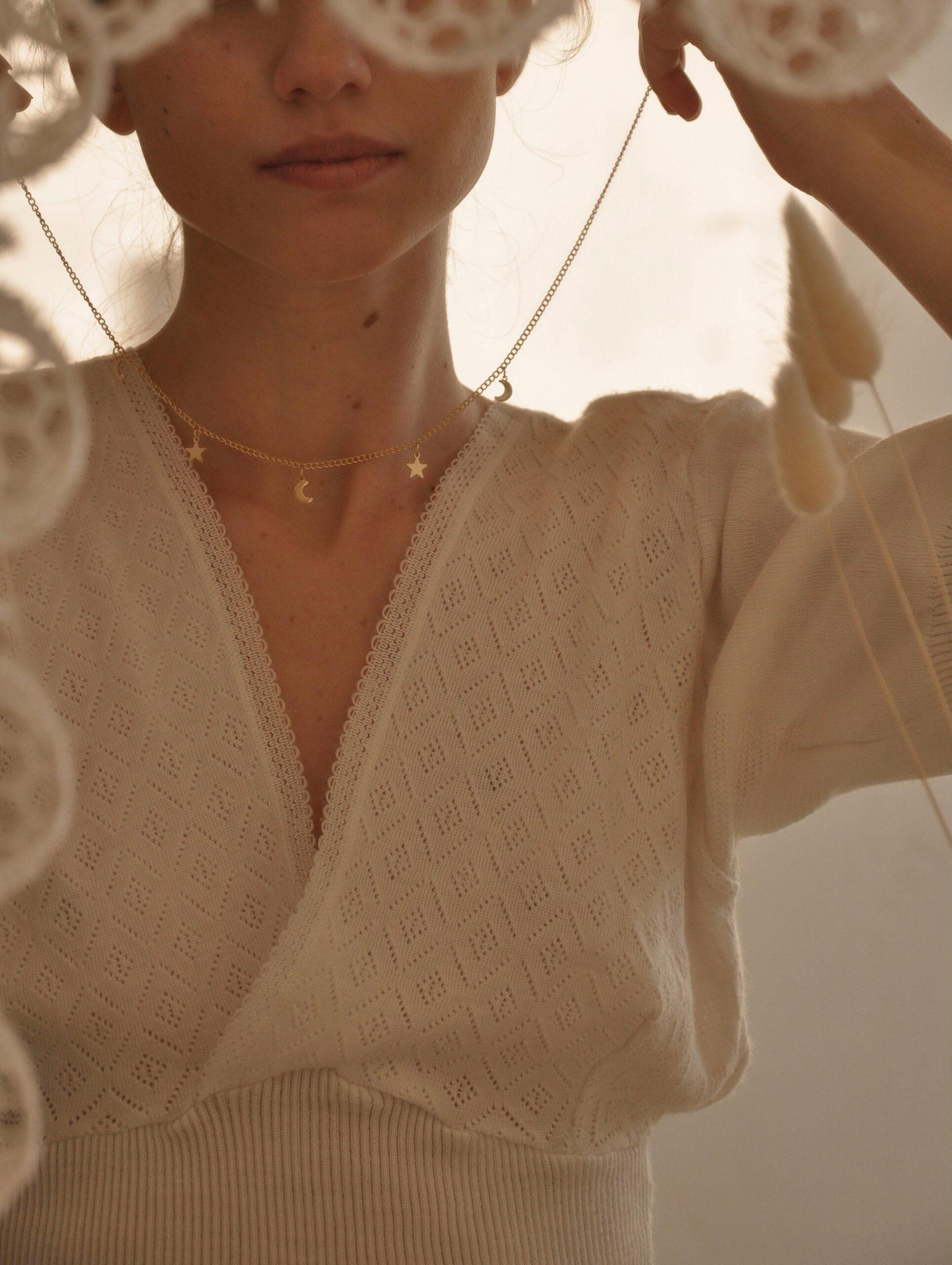 Close-up of a young woman partially concealed behind hanging lace decorations, holding a delicate gold necklace adorned with tiny star and moon charms. She wears a soft, textured cream sweater with a lace trim, capturing a serene and dreamy atmosphere.