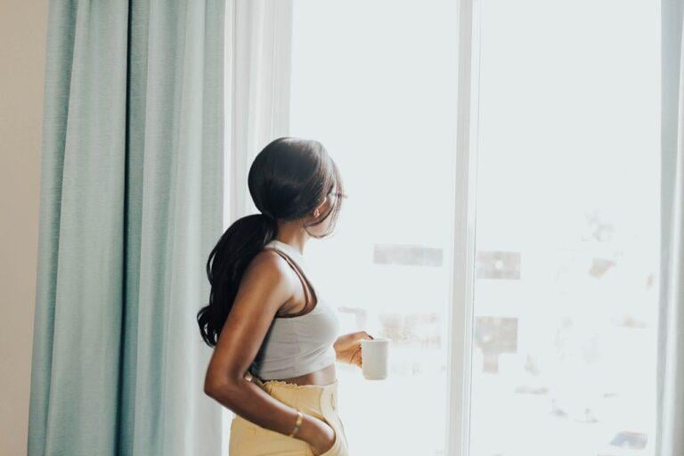A woman with a ponytail stands by a window, holding a cup of coffee and looking outside. She is dressed casually in a tank top and high-waisted pants, embodying the relaxed and peaceful vibe of a slow morning.