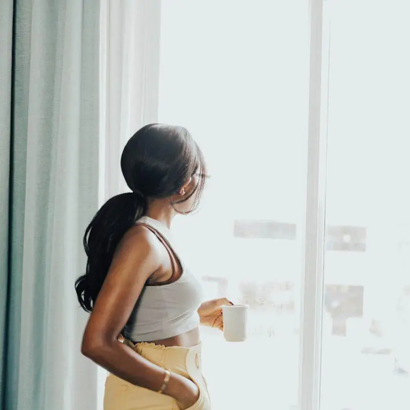 A woman with a ponytail stands by a window, holding a cup of coffee and looking outside. She is dressed casually in a tank top and high-waisted pants, embodying the relaxed and peaceful vibe of a slow morning.