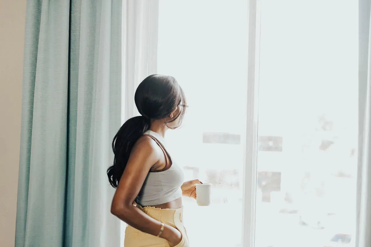 A woman with a ponytail stands by a window, holding a cup of coffee and looking outside. She is dressed casually in a tank top and high-waisted pants, embodying the relaxed and peaceful vibe of a slow morning.