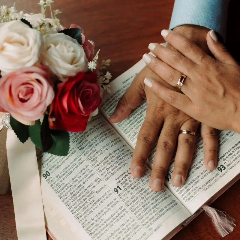 A bouquet of red, pink, and white roses rests on an open Bible, with a couple's hands, both wearing wedding rings, gently placed over the pages. This image accompanies a blog post titled "Wedding Prayers and Blessings for a Beautiful Ceremony," highlighting the theme of love and spiritual unity.