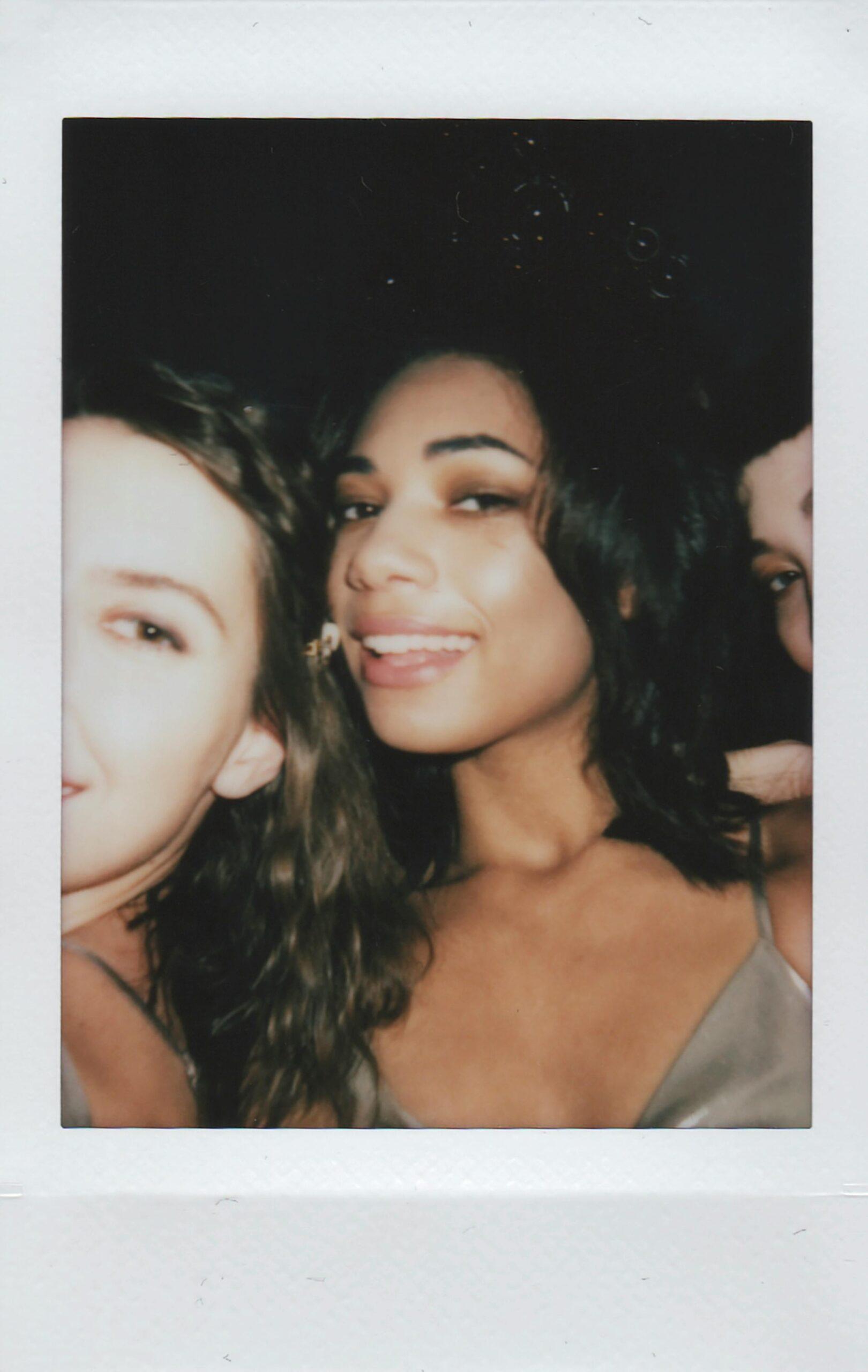A candid polaroid photo of three female friends, smiling and posing closely together at a party. The central figure playfully sticks out her tongue, adding a fun and flirty vibe to the moment.