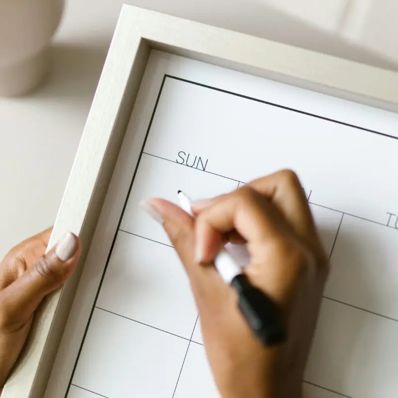 A close-up image shows a woman's hand holding a whiteboard calendar and writing on the "Sunday" section with a black marker. The image suggests the beginning of planning or organizing a schedule, possibly for a weekly or monthly challenge. The scene conveys a sense of preparation and intention-setting.