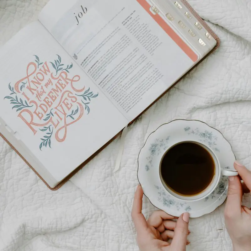 Open Bible on a white bedspread, displaying a decorative page with the words "I know that my Redeemer lives" and the start of the book of Job. Next to the Bible, a woman holds a floral teacup filled with coffee. This serene setting emphasizes peaceful reflection, ideal for a beginner's Bible reading plan.