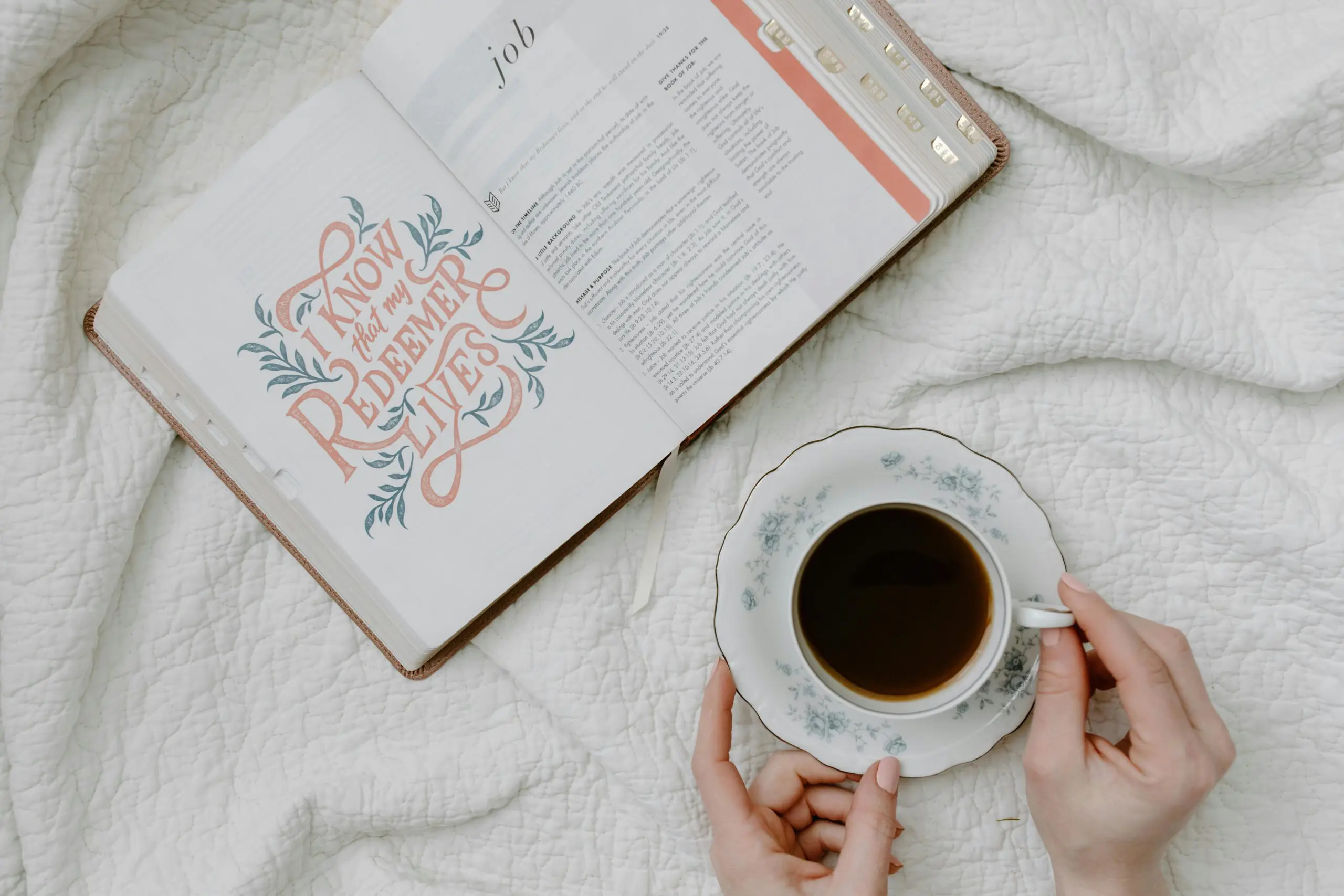 Open Bible on a white bedspread, displaying a decorative page with the words "I know that my Redeemer lives" and the start of the book of Job. Next to the Bible, a woman holds a floral teacup filled with coffee. This serene setting emphasizes peaceful reflection, ideal for a beginner's Bible reading plan.