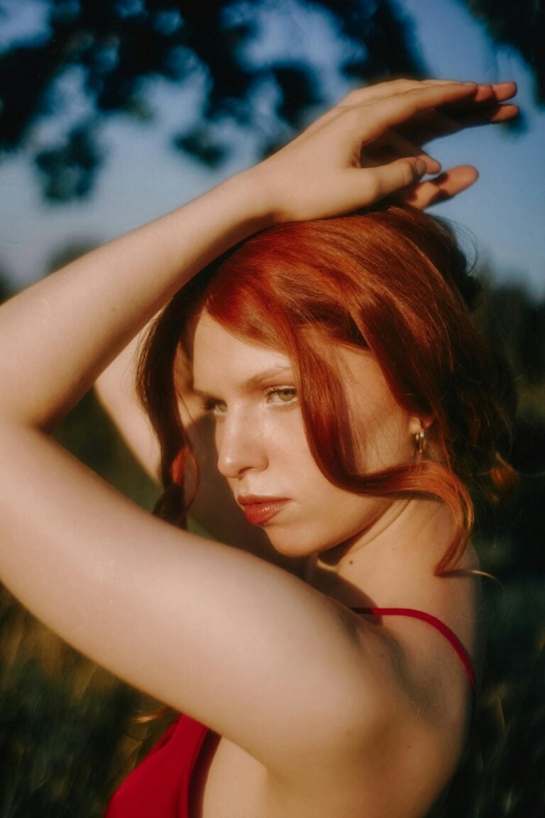 A woman with striking red hair gazes intensely off-camera, her hands gracefully positioned above her head. The warm lighting highlights her porcelain skin and the natural setting in the background, creating an air of mystery and allure.