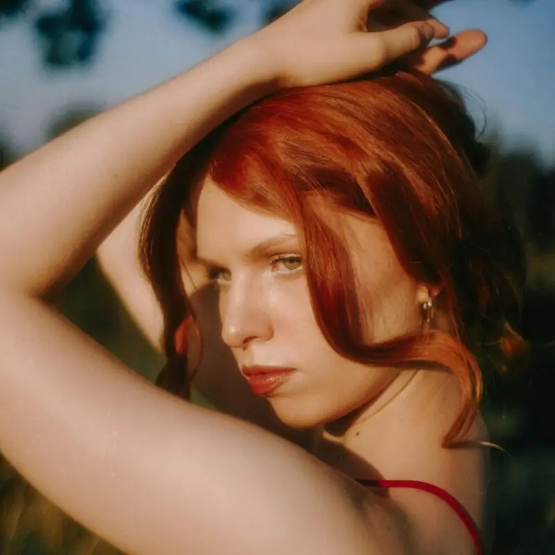 A woman with striking red hair gazes intensely off-camera, her hands gracefully positioned above her head. The warm lighting highlights her porcelain skin and the natural setting in the background, creating an air of mystery and allure.