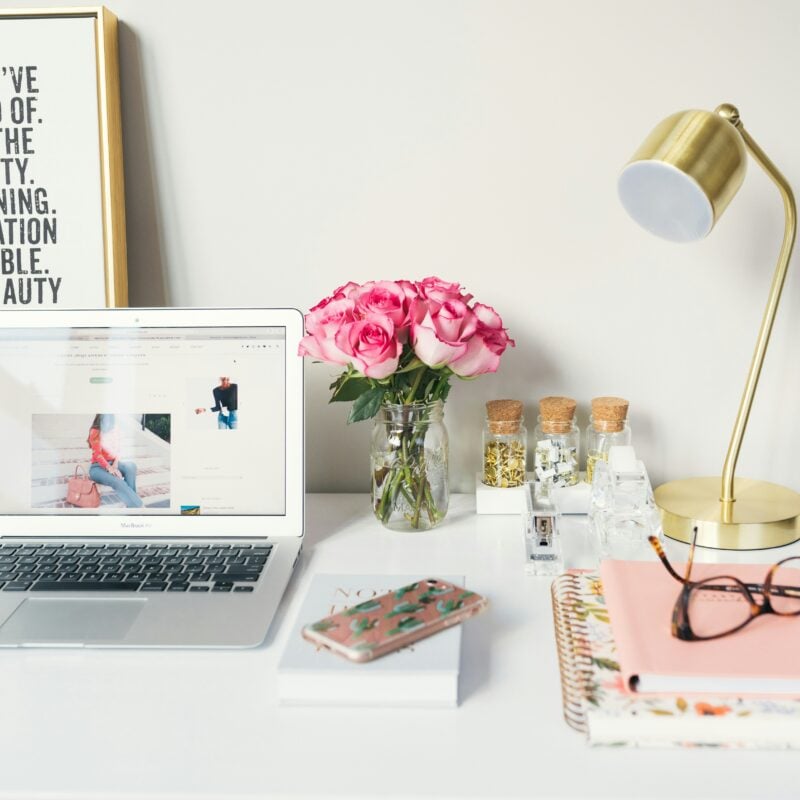 A tidy, aesthetically pleasing workspace with a MacBook displaying a blog, a framed motivational quote, a vase of pink roses, a gold desk lamp, notebooks, a smartphone, and various desk accessories, emphasizing an environment conducive to focus and productivity.