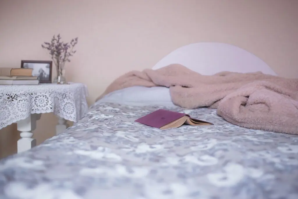 A cozy bedroom setup highlighting an essential part of an evening routine, featuring a softly textured blanket on a bed with a floral patterned duvet. A bedside table is adorned with lavender, books, and a vintage photo, creating a tranquil reading nook.