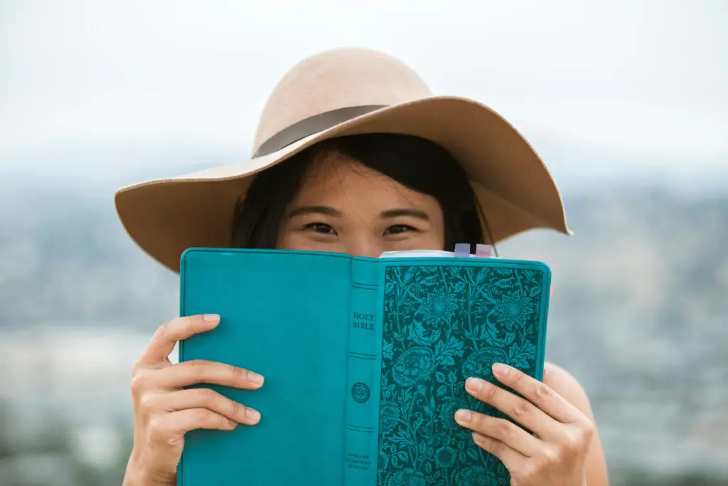 A young woman in a wide-brimmed hat holds up a turquoise Bible in front of her face, partially obscuring it. Her eyes peek over the top, hinting at a playful or contemplative mood, with a blurred background suggesting a high vantage point.