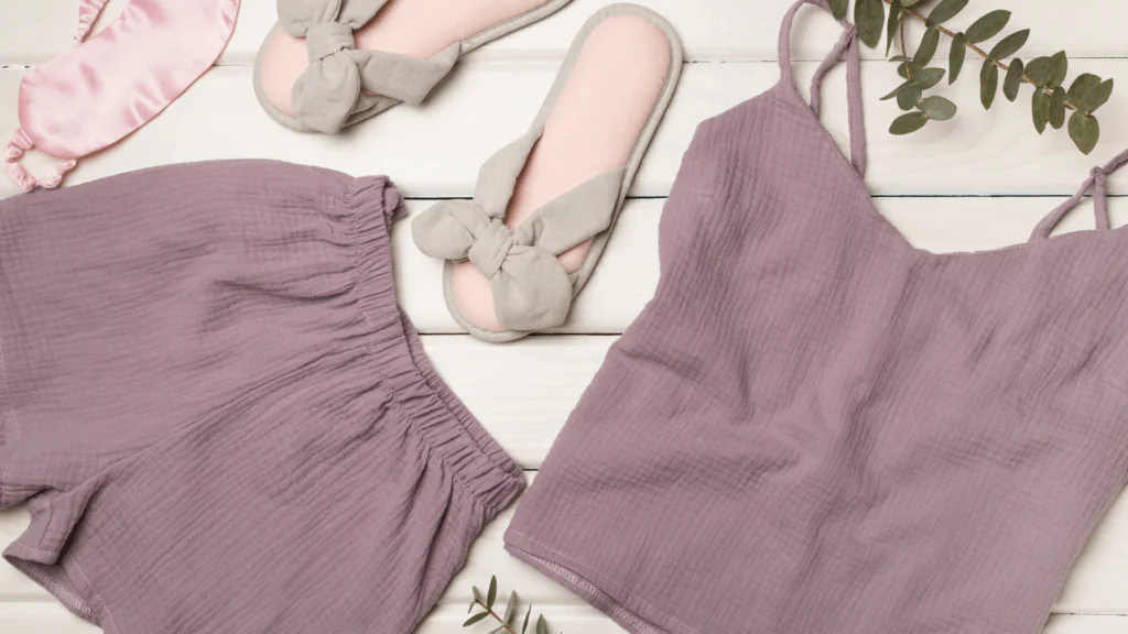 A flat lay of comfortable bedtime essentials including a purple sleep mask, soft slippers with a bow, a textured camisole top, and crinkled pajama shorts. Green leaves subtly decorate the arrangement, promoting a restful evening routine atmosphere.