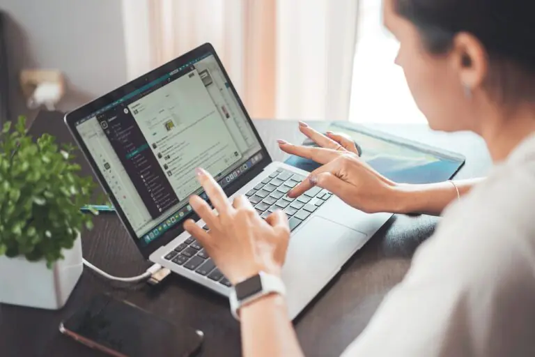 A woman in a business casual outfit uses her laptop while sitting at a wooden desk, surrounded by a vibrant green plant, a smartphone, and various digital devices. The screen displays multiple communication apps, showcasing a modern, organized workspace ideal for smart working.