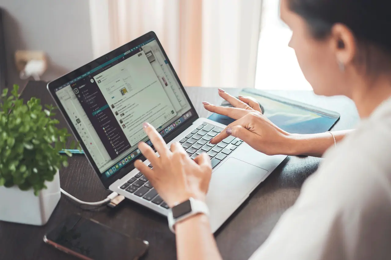 A woman in a business casual outfit uses her laptop while sitting at a wooden desk, surrounded by a vibrant green plant, a smartphone, and various digital devices. The screen displays multiple communication apps, showcasing a modern, organized workspace ideal for smart working.