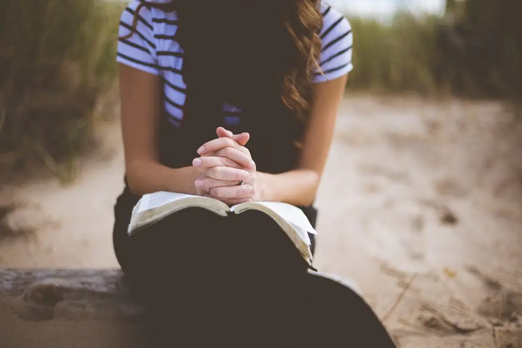 A young woman sits outdoors on a wooden bench, her hands clasped in prayer over an open Bible resting on her lap. The setting is serene, with a blurred sandy path and greenery in the background, capturing a moment of peaceful devotion.