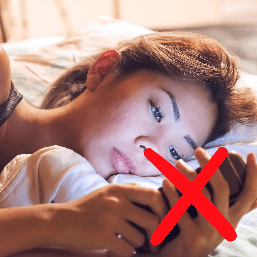 A woman lying in bed looks intently at her smartphone, symbolizing a common habit to avoid when establishing a beneficial evening routine. The red cross over the image underscores the negative impact of screen time before sleep.