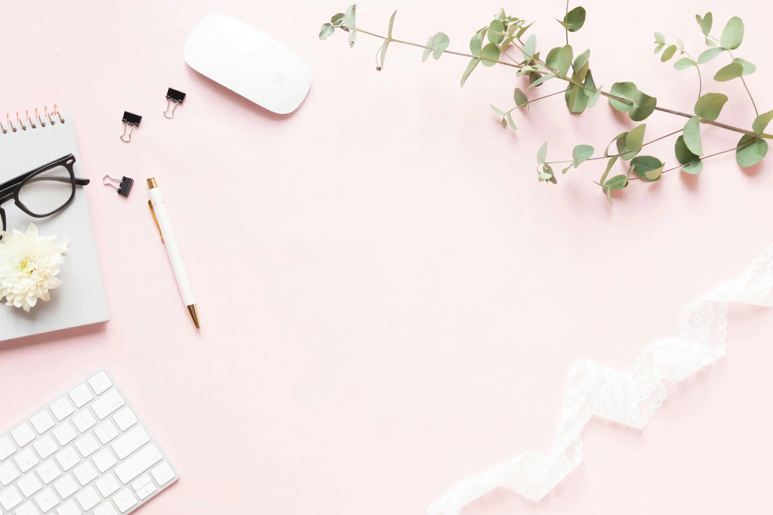 A flat lay workspace on a soft pink background featuring a grey notebook with a white flower on it, a white pen, a keyboard, and a computer mouse. Decorative elements include black binder clips, a sprig of green eucalyptus, and a delicate white lace ribbon, creating an elegant and creative setting.