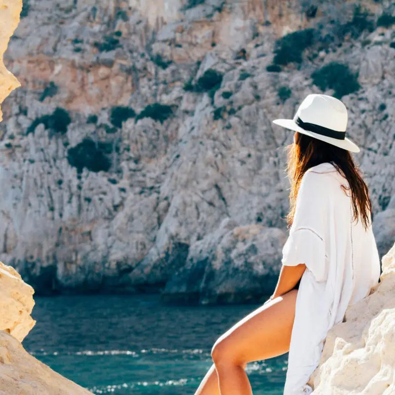 A woman in a white cover-up and a wide-brimmed hat sits on a rocky ledge, gazing out at the blue sea and rugged cliffs. The serene coastal setting reflects a sense of adventure and tranquility, perfect for capturing the essence of travel and trip captions.