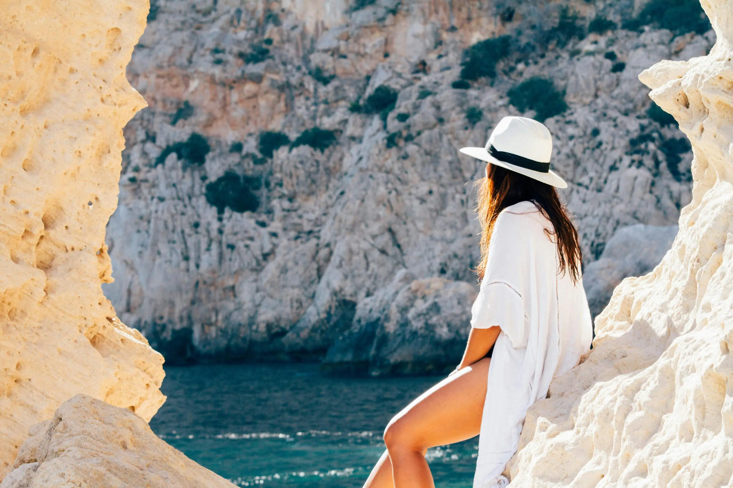 A woman in a white cover-up and a wide-brimmed hat sits on a rocky ledge, gazing out at the blue sea and rugged cliffs. The serene coastal setting reflects a sense of adventure and tranquility, perfect for capturing the essence of travel and trip captions.