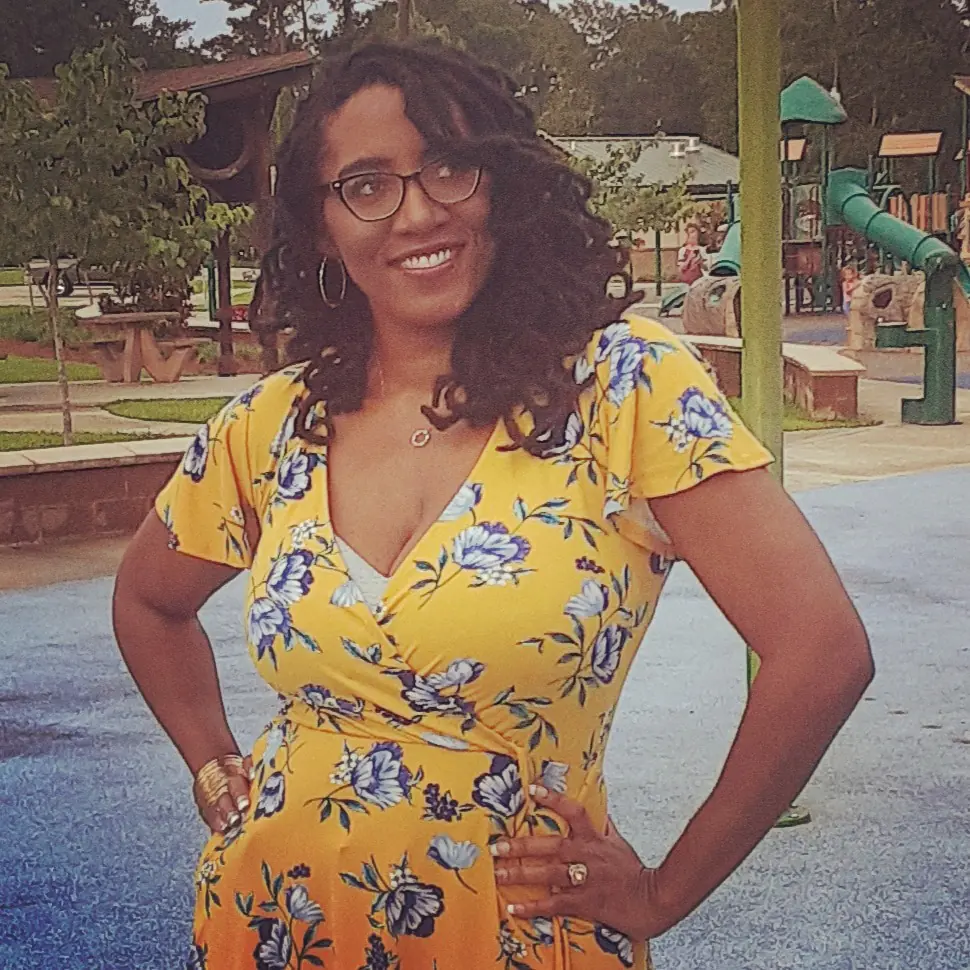 LaToya Rachelle, smiling warmly, stands in a park with playground equipment in the background. She wears a vibrant yellow wrap dress adorned with a blue floral pattern, accessorized with hoop earrings, glasses, and a heart-shaped necklace. Her curly hair frames her face, adding a friendly and approachable look to her author bio.