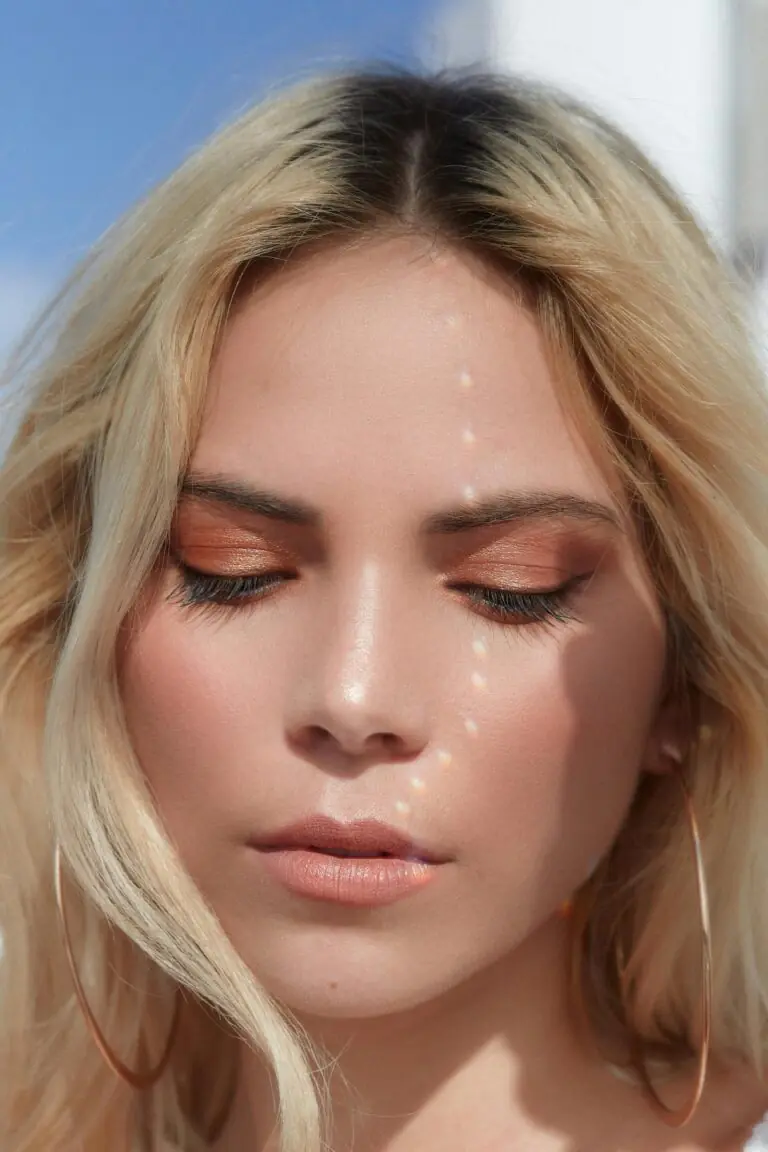 This is a close-up image of a woman with her eyes closed, wearing peach-toned eyeshadow and minimal makeup. Light reflections form a dotted line across her face, creating a soft, artistic effect. Her blonde hair frames her face, and she wears large hoop earrings.