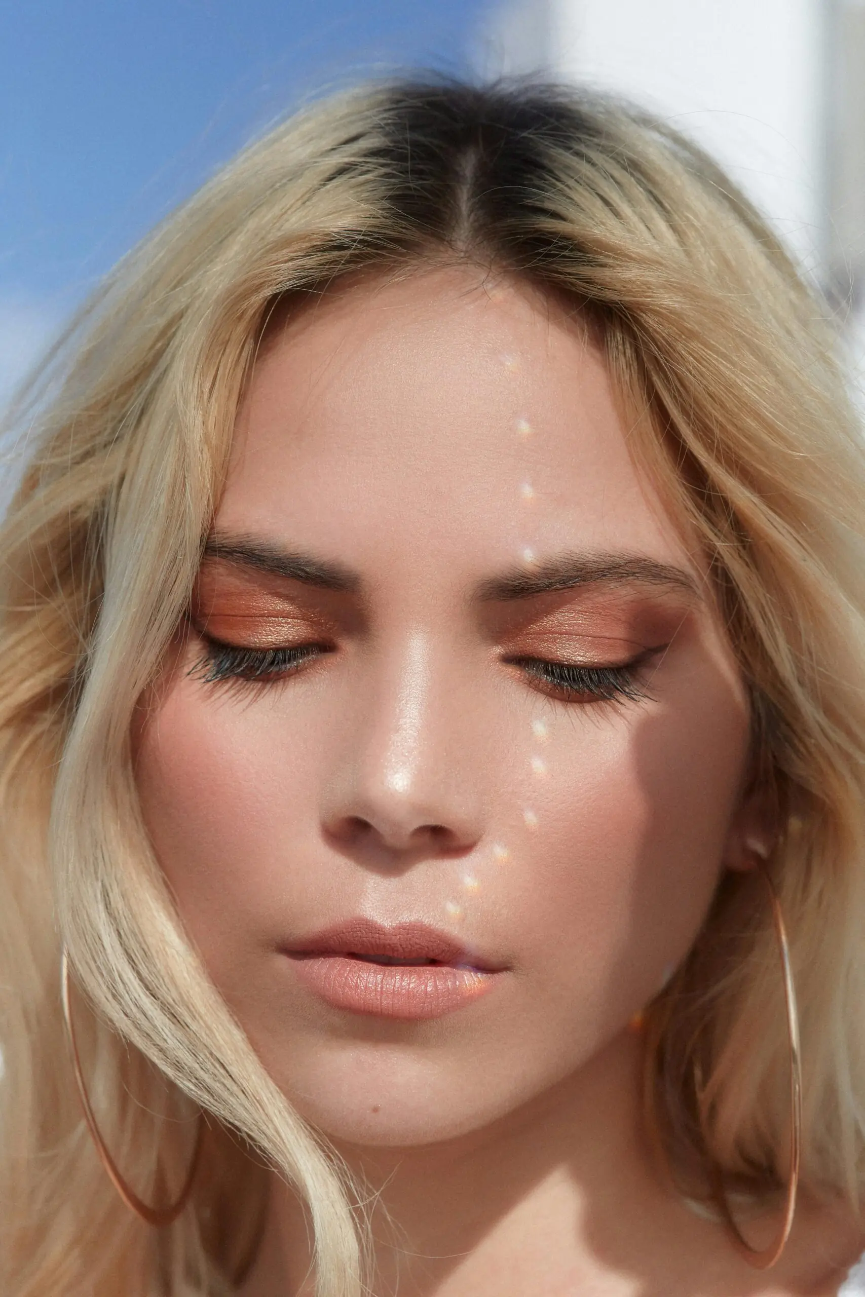 This is a close-up image of a woman with her eyes closed, wearing peach-toned eyeshadow and minimal makeup. Light reflections form a dotted line across her face, creating a soft, artistic effect. Her blonde hair frames her face, and she wears large hoop earrings.