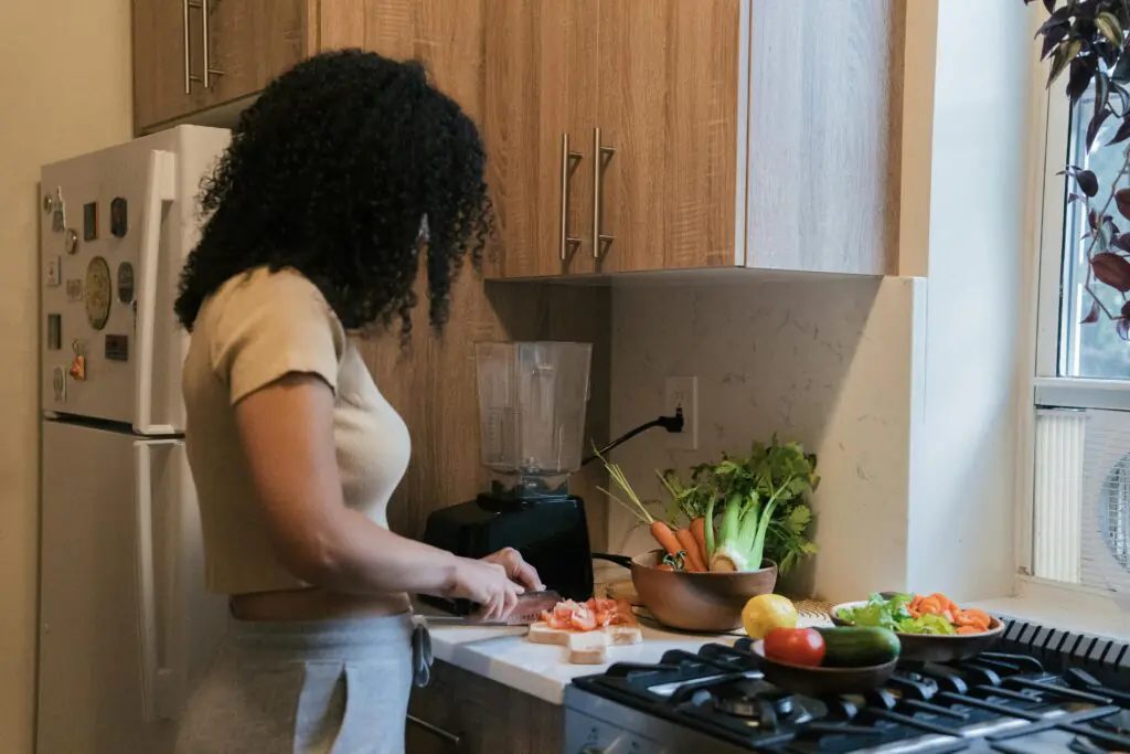 A woman preparing food in a kitchen, chopping tomatoes on a cutting board surrounded by fresh vegetables like carrots, celery, and cucumbers, emphasizing healthy home cooking.