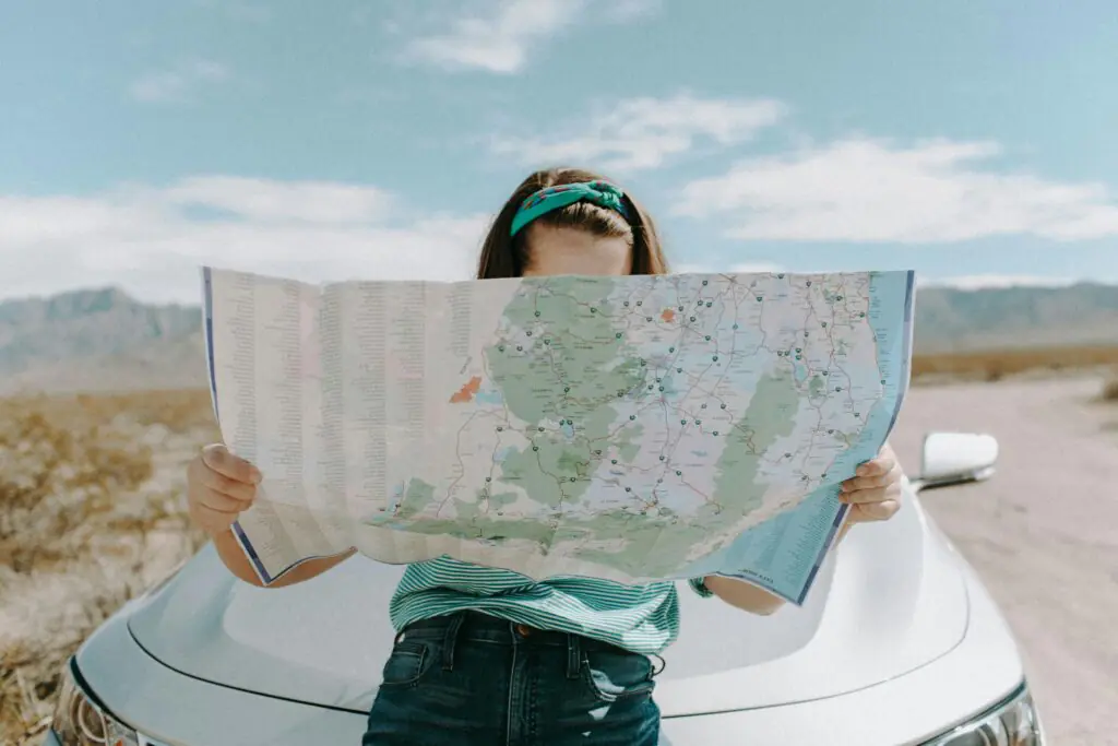 A woman leaning against a car in a scenic, open landscape, holding a large map to navigate, symbolizing the spirit of travel and exploring new places.