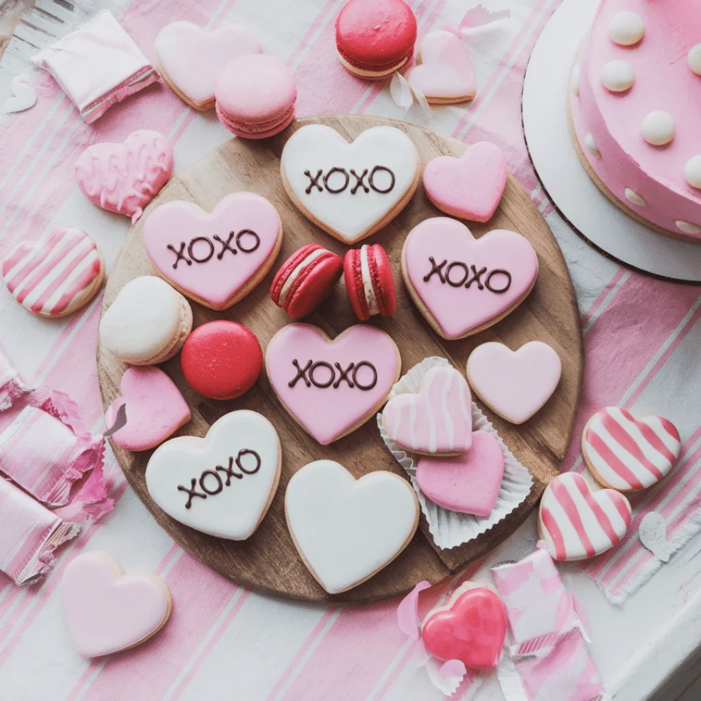 A flat-lay of a wooden tray filled with heart-shaped cookies decorated in pink, white, and red, some with "XOXO" written on them. Surrounding the tray are colorful macarons, striped and frosted cookies, and a pink polka-dotted cake on the side. The scene is festive and sweet, perfect for a Galentine's Day celebration.