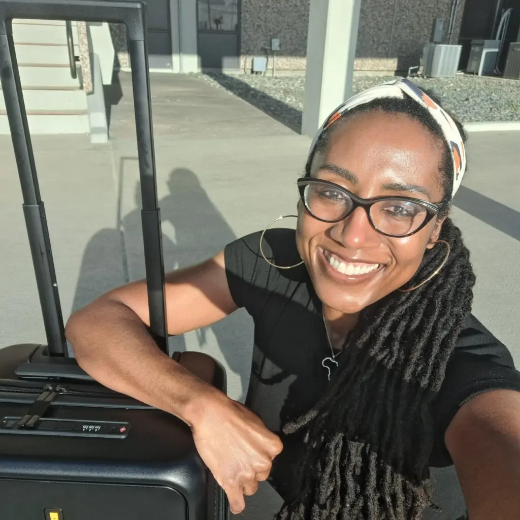 A joyful woman poses for a sunny selfie while leaning on a black carry-on suitcase with an extended handle. She’s wearing a black top, large hoop earrings, glasses, and a headband over her long locs, along with a necklace featuring an Africa-shaped pendant. The background includes a modern building with steps and a concrete walkway.