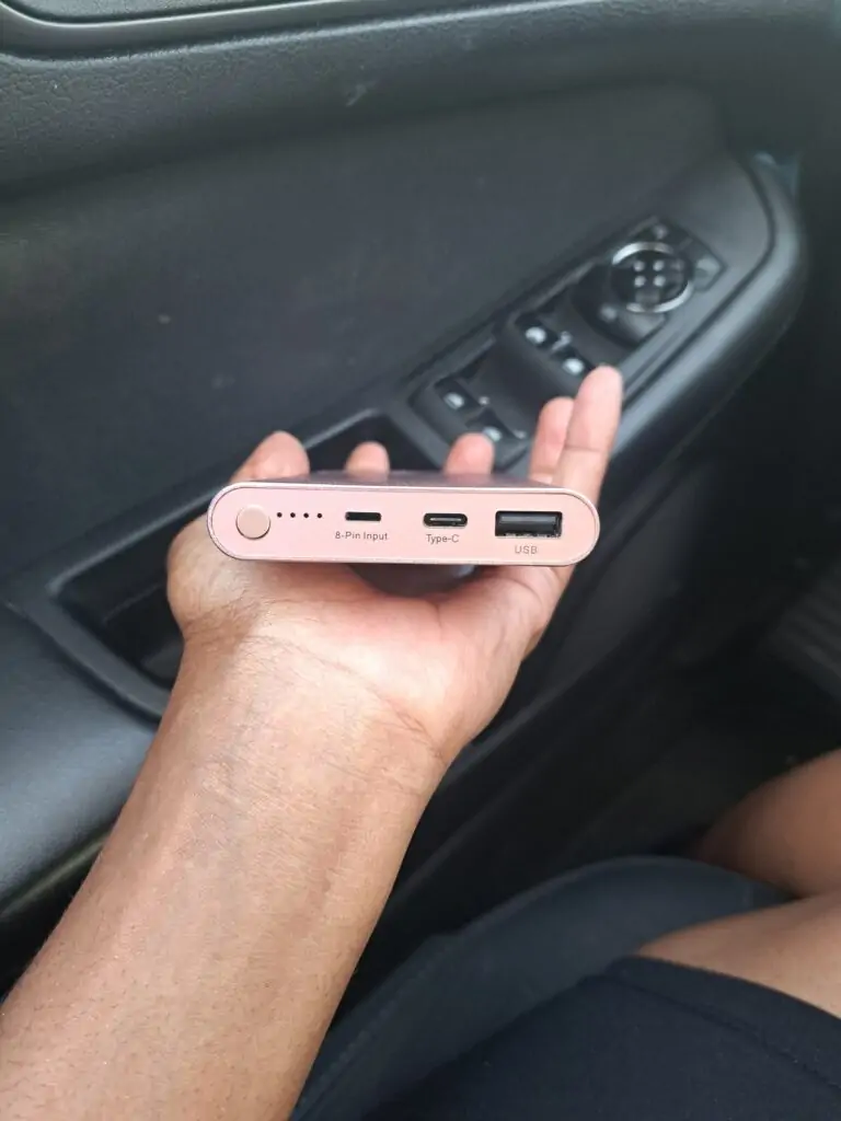 A person holds a slim, rose gold portable charger in a car, showing its ports labeled 8-Pin Input, Type-C, and USB along with LED indicator lights and a power button. The charger rests in their palm with a black car door and window controls visible in the background.