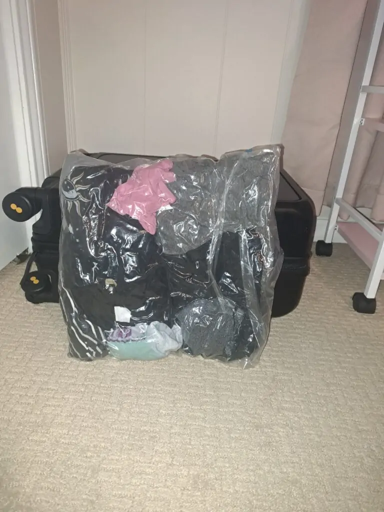 A fully compressed vacuum-seal storage bag containing various clothing items, including a pink garment, striped and celestial-patterned pieces, and rolled socks. The bag stands upright on a beige carpet in front of a closed black suitcase, visibly reduced in size after air has been removed.