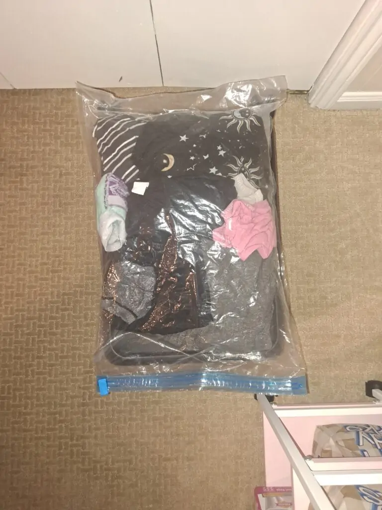 A large clear vacuum-seal storage bag filled with various folded and rolled clothing items, including black, pink, gray, and striped garments. The bag is uncompressed, sealed at the bottom with a blue zipper closure, and placed on a beige carpeted floor near a white door.