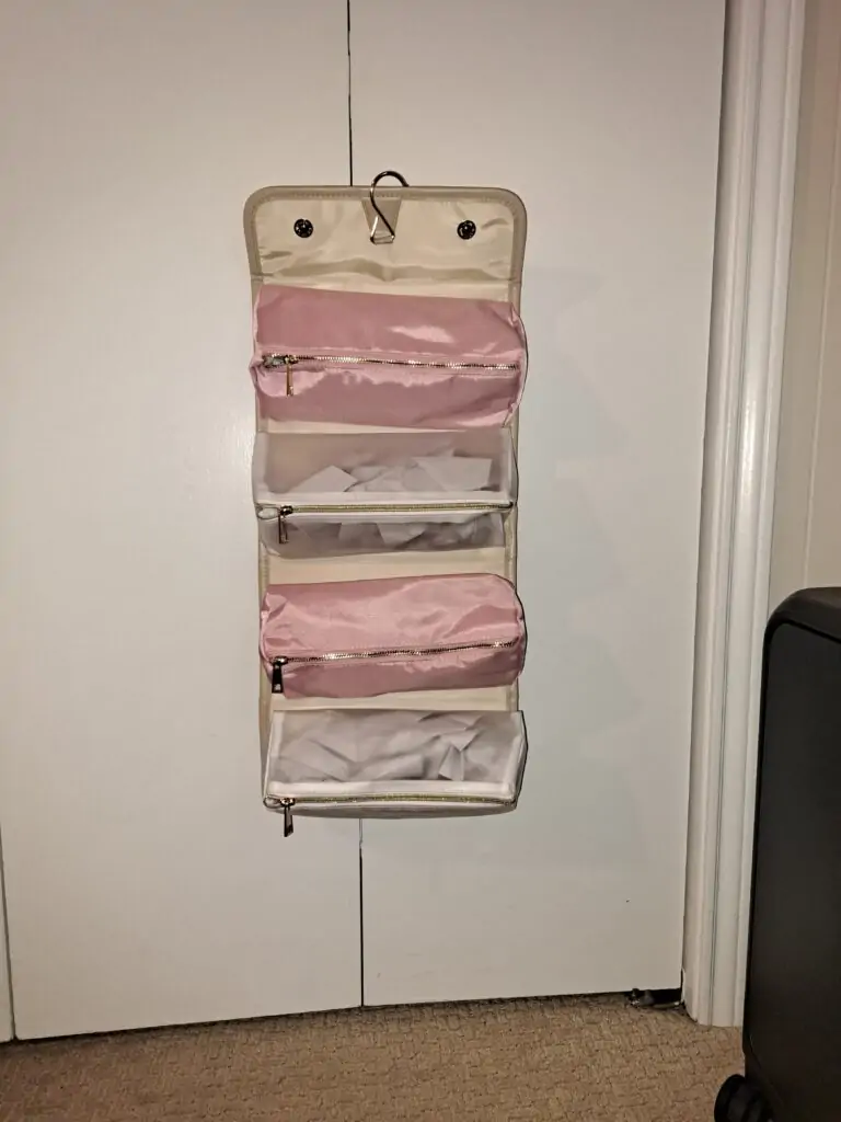 A hanging roll-up travel organizer with a beige exterior and multiple zippered compartments, including pink and clear sections for easy visibility of contents. It’s displayed hanging on a white door using a metal hook, with a black suitcase partially visible to the side.